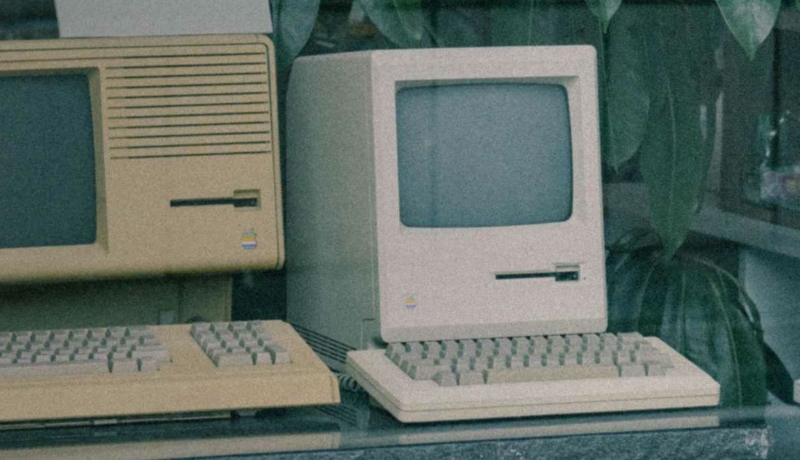 Dwa retro komputery Apple – Macintosh oraz Apple II – stojące obok siebie na blacie w otoczeniu roślin doniczkowych.