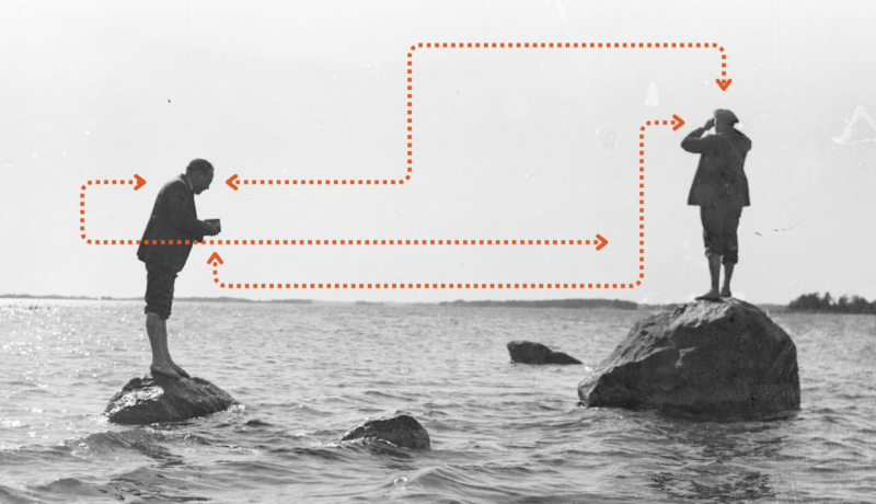 Archiwalne, czarno-białe zdjęcie dwóch mężczyzn stojących na głazach w morzu. Jeden z nich patrzy przez lornetkę w dal, drugi robi mu zdjęcie.