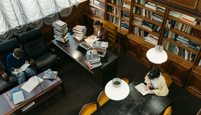 Wnętrze biblioteki – trzy osoby siedzą przy stołach, pracują z wykorzystaniem wielu książek.